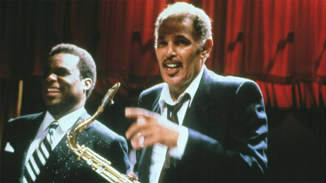 A man stands on stage talking and pointing. He holds a saxophone. Next to him is another man in a suit, laughing.