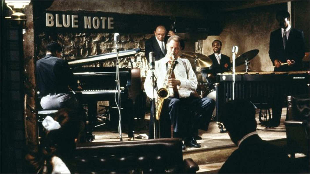 A man in a white shirt and overalls is seated and playing a saxophone on a stage in the Blue Note. He is surrounded by a piano player, a person playing the vibraphone, a drummer, and a person playing upright bass.