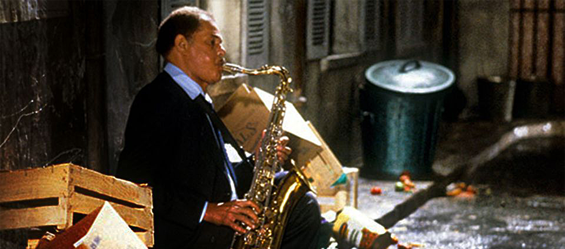 A man, seated in an alley plays a saxophone. He is surrounded by boxes and garbage.