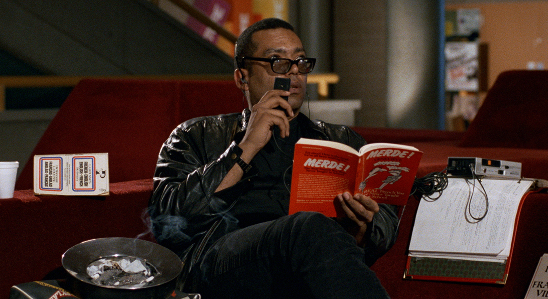 A man in eyeglasses sits on the couch. In one hand is an open book and in the other he holds a small tape recorder into which he speaks.