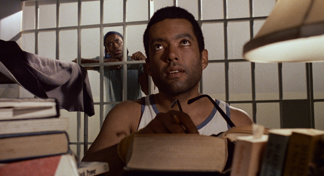 A man in a jail cel sits at a small desk on which books are scattered and some are stacked. A lamp is to his left. He holds eye glasses in his hands as he looks upward at another person not in frame