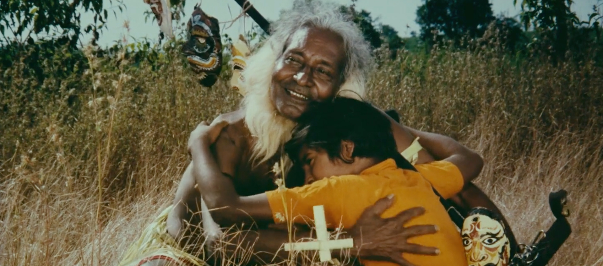 An elder with white hair smiles and holds a young child in a yellow shirt. A cross sits in front of them as they both sit in a field