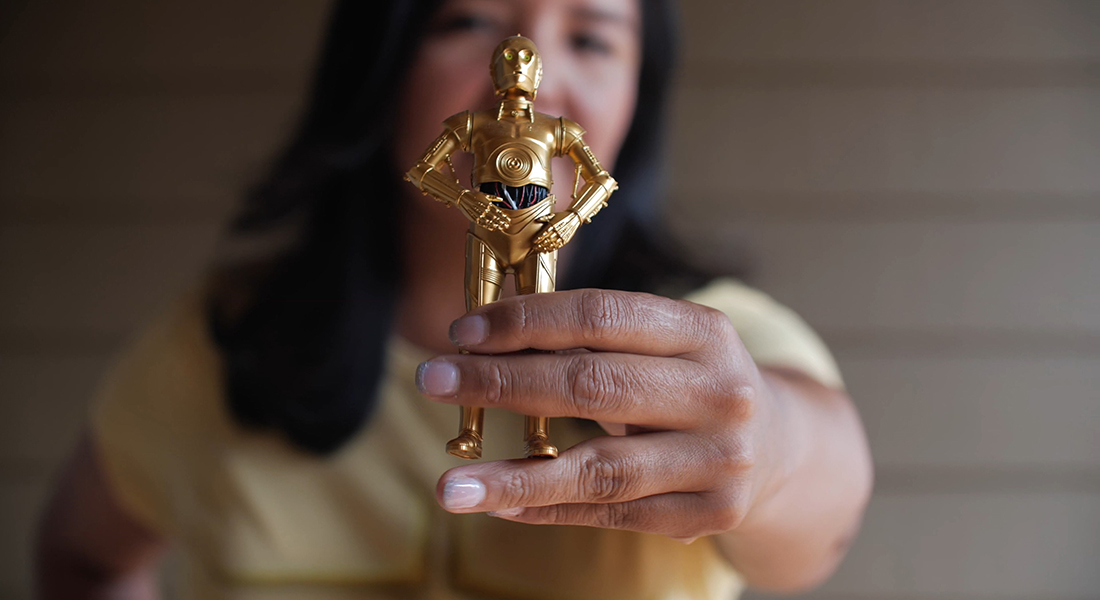 A close-up of a C-3PO action figure which is gold and has its arms on its hips. It is held by a person who is in the background