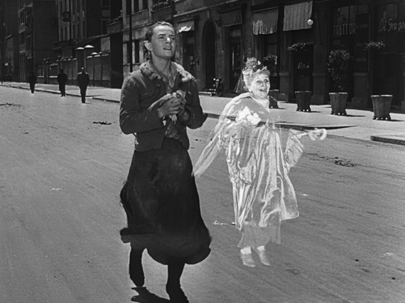 A woman in a tattered dress walks down an empty street alongside the ghostly figure of an older woman