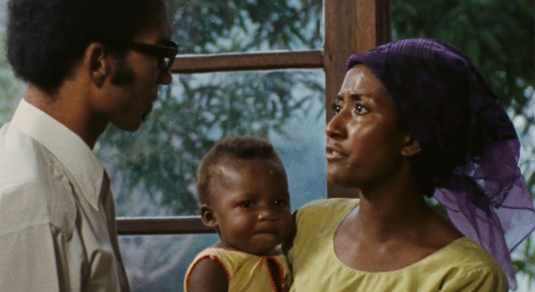 A woman in a purple headdress and yellow shirt hold a baby and she stares intensely at a man in glasses and a white shirt. Behind them both is a window looking out to leaves and trees