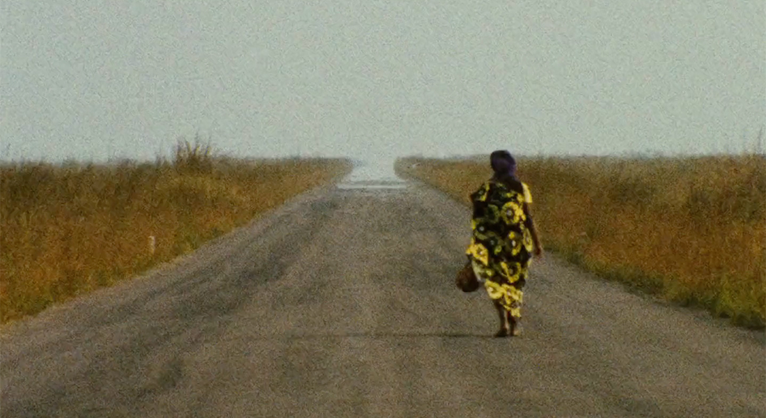 A woman in colorful clothes walks down a wide, empty road. On both sides of the road is dry brush