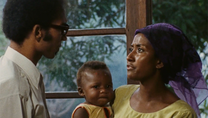 A woman in a purple headdress and yellow shirt hold a baby and she stares intensely at a man in glasses and a white shirt. Behind them both is a window looking out to leaves and trees