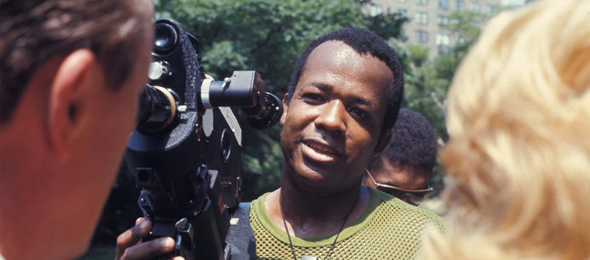 Filmmaker William Greaves stands in front of a man and a woman. He is holding a film camera.