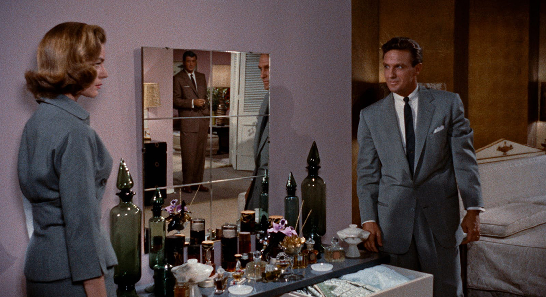Separated by a drink cart and mirror reflecting a man in a doorway, a man in a gray suit stares down a woman in a gray dress