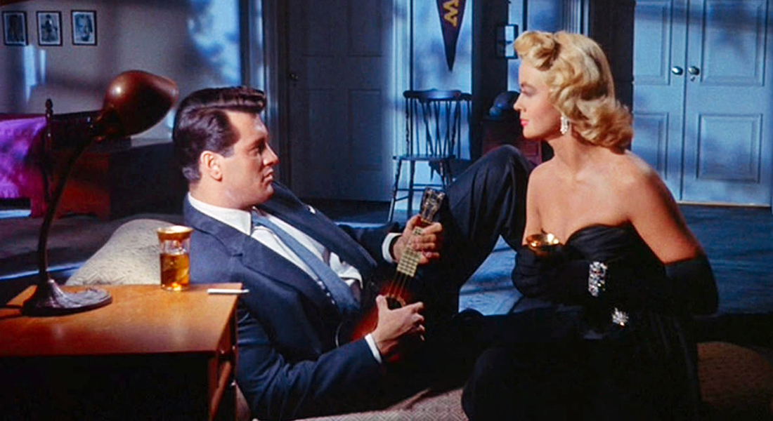 A man in a blue suit lounges on a chaise lounge. He holds a ukelele and is making eyes and a blond women in a dark gown