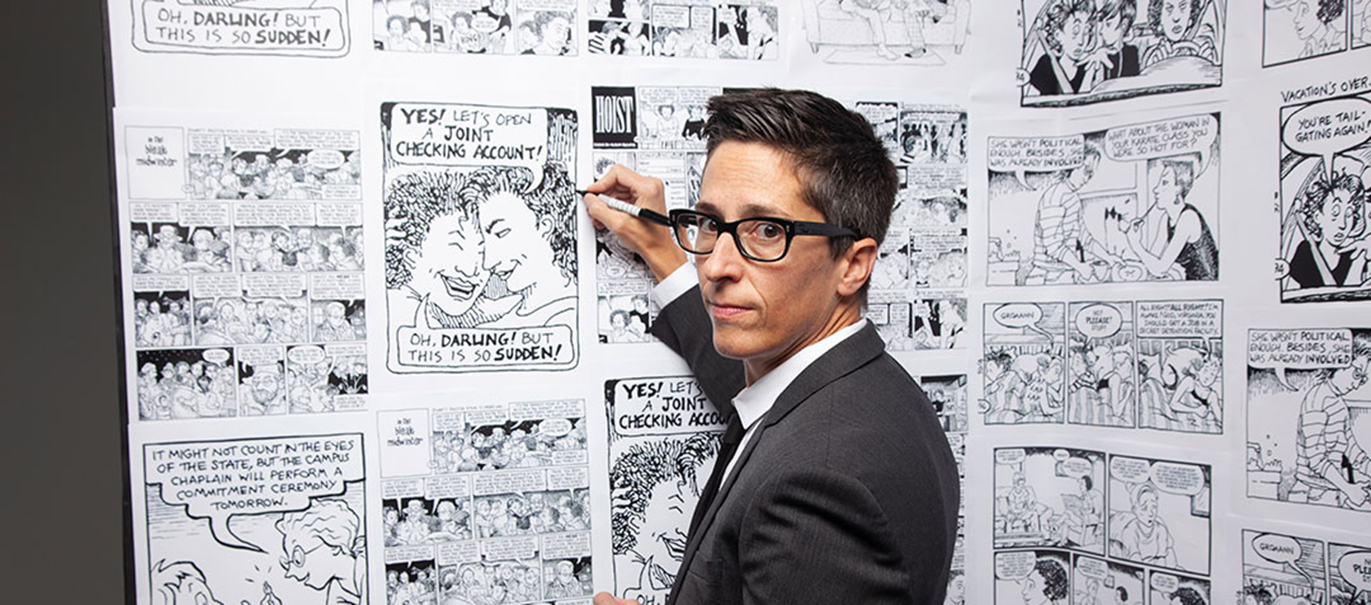 Photo of cartoonist Alison Bechdel looking over her shoulder at the camera. She has short, dark hair and is wearing a suit and glasses. She is holding a marker against a white backdrop full of comics in black ink. 