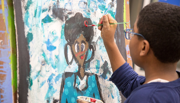 Three-quarter profile view of young black boy with glasses as he paints a large, colorful portrait on an easel