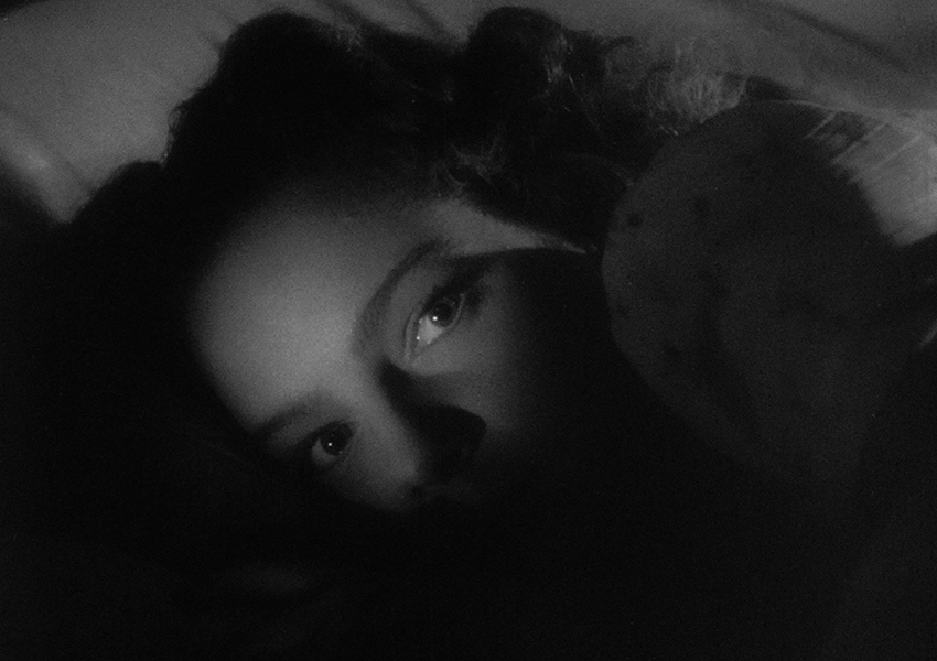 Black-and-white still of a person’s face lit up in the dark; they appear to be laying down in bed. 