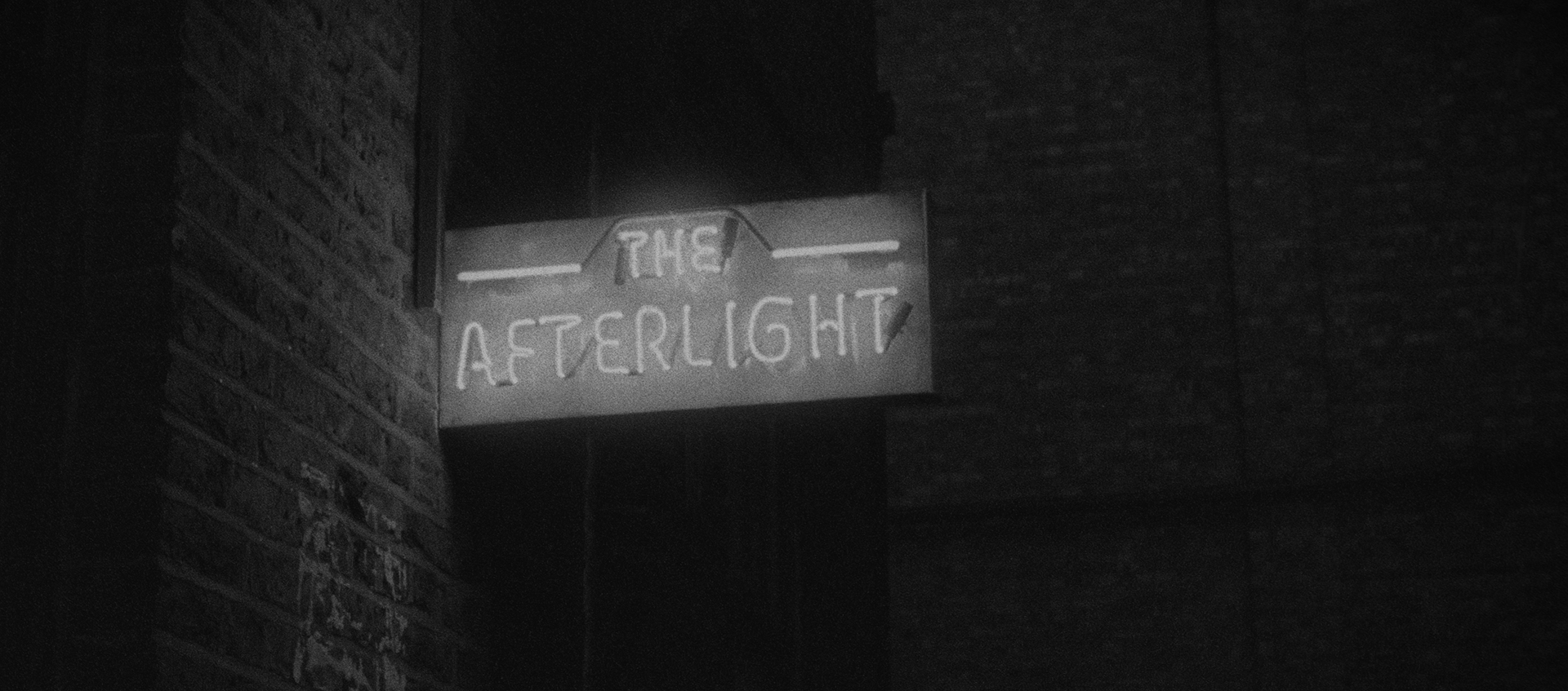 Black-and-white still of a neon-lit sign that says The Afterlight; it is protruding from a brick wall.
