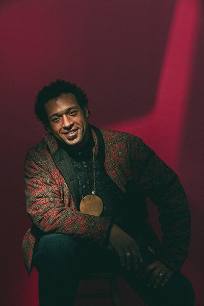 Makaya McCraven, who has dark hair and is wearing a large golden circle pendant and a patterned red jacket, sits in front of a red background; he is smiling and has his right arm on one knee and his left hand on the other. 