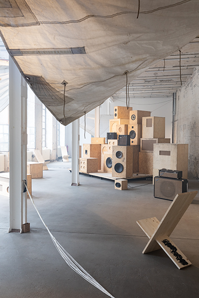 Installation view of nora chipaumire's afternow, featuring the monumental speaker system soundshitsystem, which has multisized wooden speakers stacked on top of each other in the corner of a gallery space that has chipped, white-painted brick walls and tall, open windows on the left side of the room.
