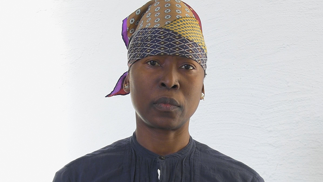 Headshot of artist nora chipaumire against a white background. She has dark brown skin and is wearing a multicolored, patterned headscarf and a dark, button-up shirt. She gazes directly into the camera.