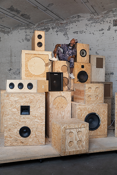 Artist nora chipaumire sits atop soundshitsystem, a tower of box-shaped speakers mostly made of raw plywood and particle board, which is part of her installation afternow. She wears a black-and-white patterned top, matching pants, white sneakers, and a black-and-white head covering. The tower of speakers sits on a wooden platform against a wall of white distressed bricks.