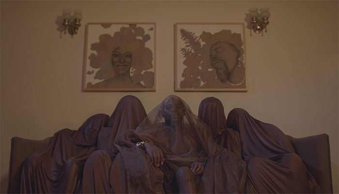 Five figures, each of whom are draped in brown fabric, sit on a brown couch. The person in the center is draped in transparent brown fabric, revealing their face and dark brown skin. Their hands, which are uncovered, rest on their legs, and there are many shiny bracelets on both wrists. Above the figures, on the wall behind them, are two portraits of Black people, both of whom have flowers and leaves surrounding their heads like hair.