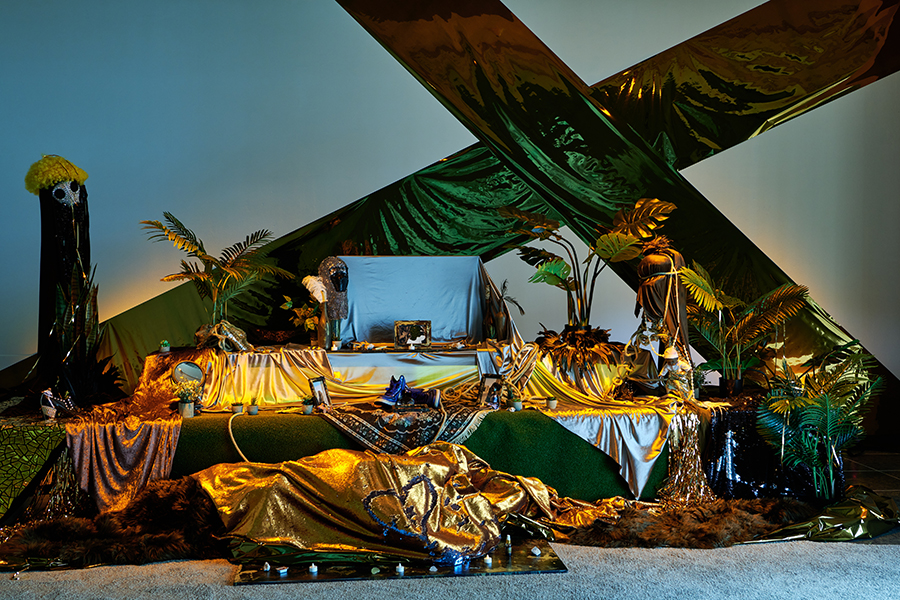 A multileveled altar draped in gold and green fabrics, featuring miscellaneous objects including a bedazzled gas mask with a curly yellow wig on top, green leafy plants, blue sneakers, a chainmail headdress, and a baby doll.