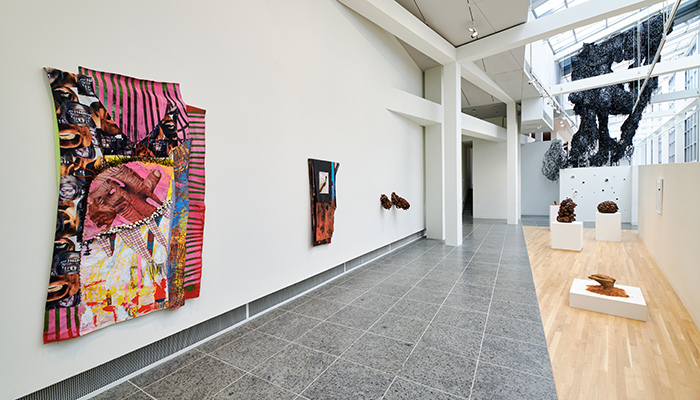 Gallery space featuring an installation of collaged, multicolored, and multilayered tapestries hanging from the white wall on the left, along with an earthwork sculpture protruding from the wall, and more resting on white pedestals on the wooden half of the gallery floor on the right. In the background, hanging from the ceiling in between the white beams of the Wex grid, is a sculpture made of VHS video tape strings and blue light.