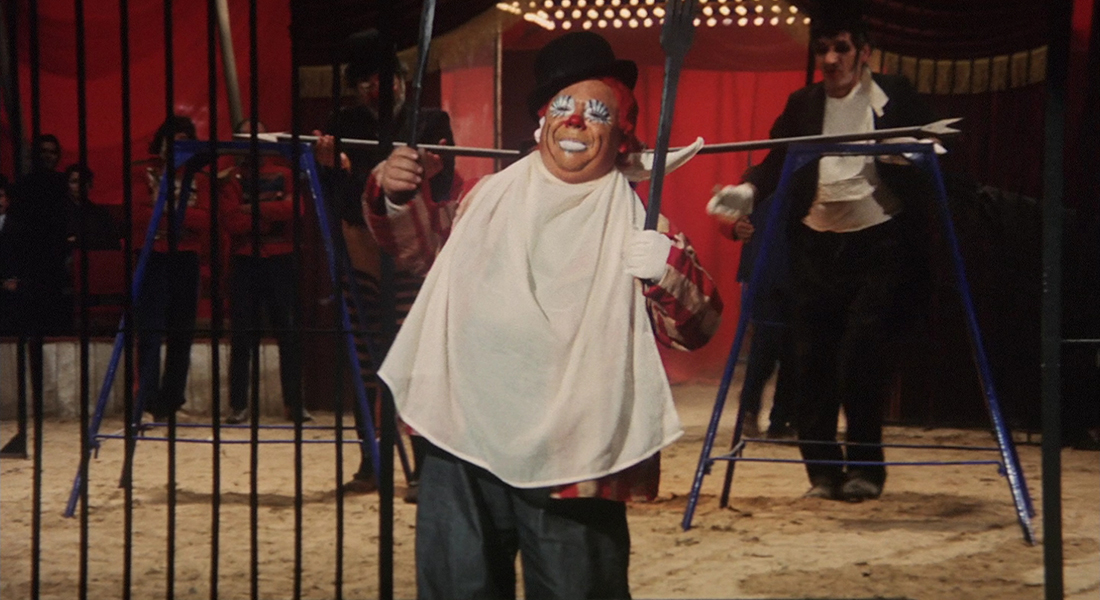 Still from I clowns featuring a person wearing a clown mask, a black hat, and a white cloth tied around their neck. They are holding metal rods in each hand, and there are metal bars to their left, with people behind them in the background in front of red curtains. The floor is covered in sand.