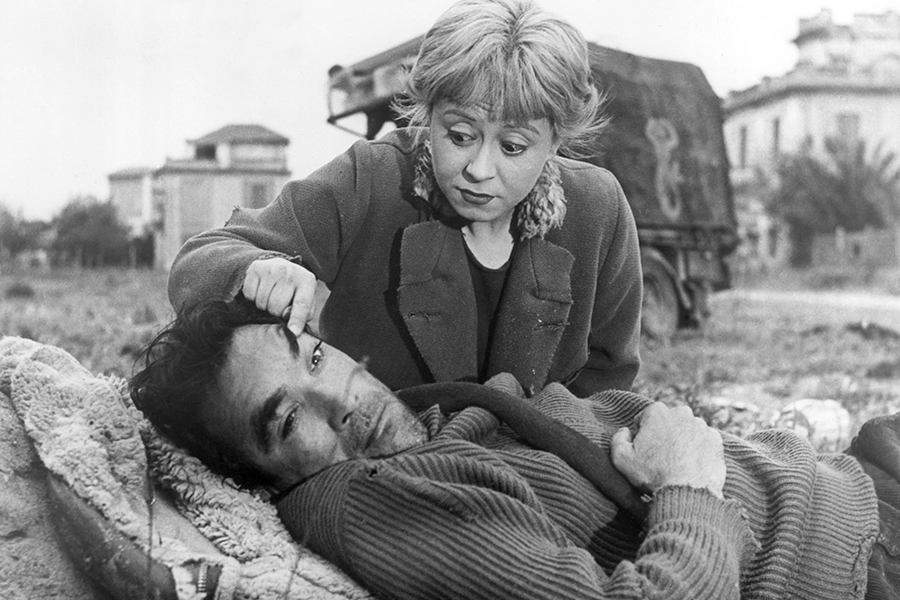 Black-and-white still of Gelsomina (Giulietta Masina) and Zampanò (Anthony Quinn) in a field. Zampanò is lying on the ground, and Gelsomina is crouched behind him, pulling his left eye open with her forefinger with a worried look on her face.