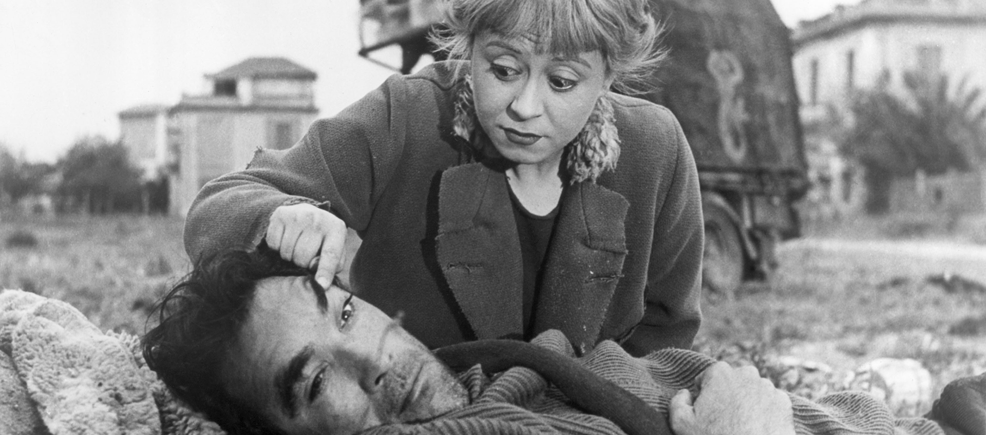 Black-and-white still of Gelsomina (Giulietta Masina) and Zampanò (Anthony Quinn) in a field. Zampanò is lying on the ground, and Gelsomina is crouched behind him, pulling his left eye open with her forefinger with a worried look on her face.