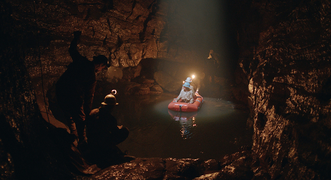 Four people at the bottom of a cave. Two are in an orange raft in a small pool, and they are lit by a spotlight from above. The other two are in the foreground on rock at the edge of the water.