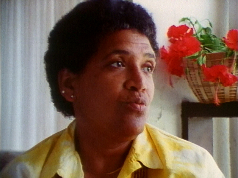 Close-up of Audre Lorde's as they look off to the side. They wear a yellow shirt and behind them is a vase of red flowers on a table