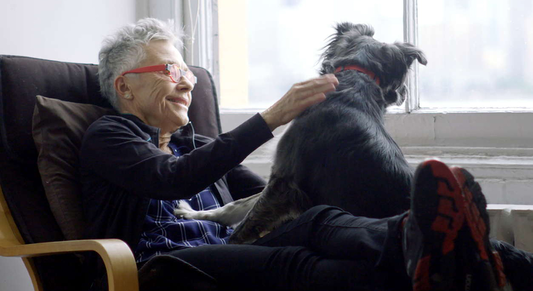 Barbara Hammer seated in a brown chair, her legs outstretched. She has short, white-gray hair and is wearing red glasses. A gray dog sits on her lap.