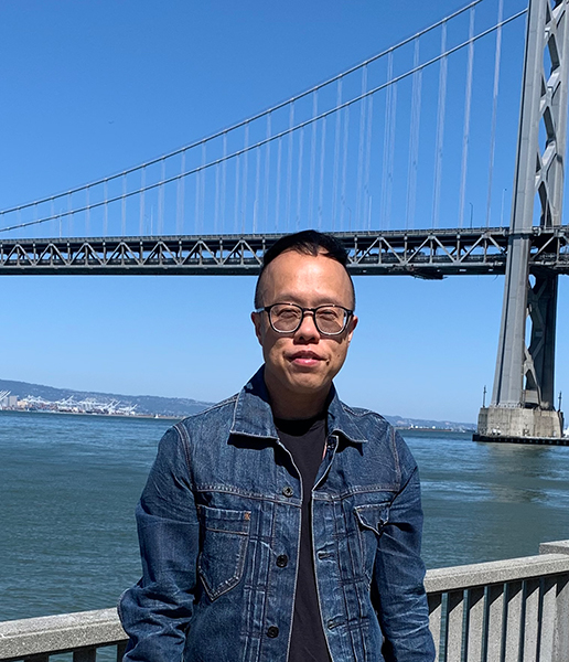 Jian Chen, who has short black hair and is wearing glasses and a denim jacket, standing in front of a bridge with a body of gray-green water underneath. There are boats and mountains on the horizon and a clear blue sky above them in the background.
