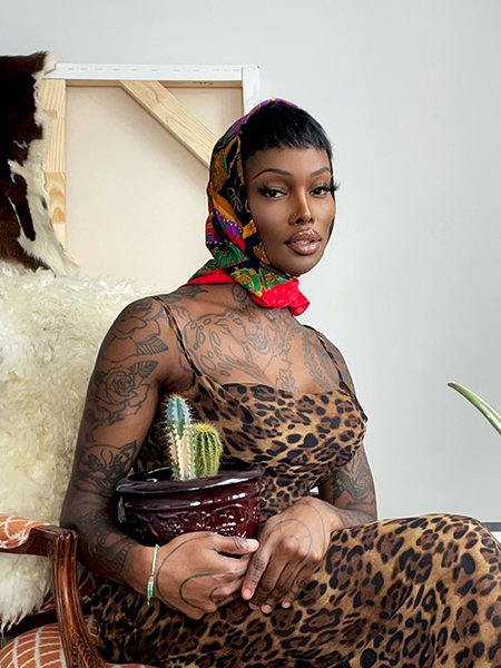 Imma Asher, who is sitting in a chair and holding a pot with two cacti in it. She is wearing a long, spaghetti-strap, cheetah-print dress, along with a multicolored, patterned headscarf. Her arms, chest, and hands are covered in black-ink tattoos featuring flowers and wild cats.