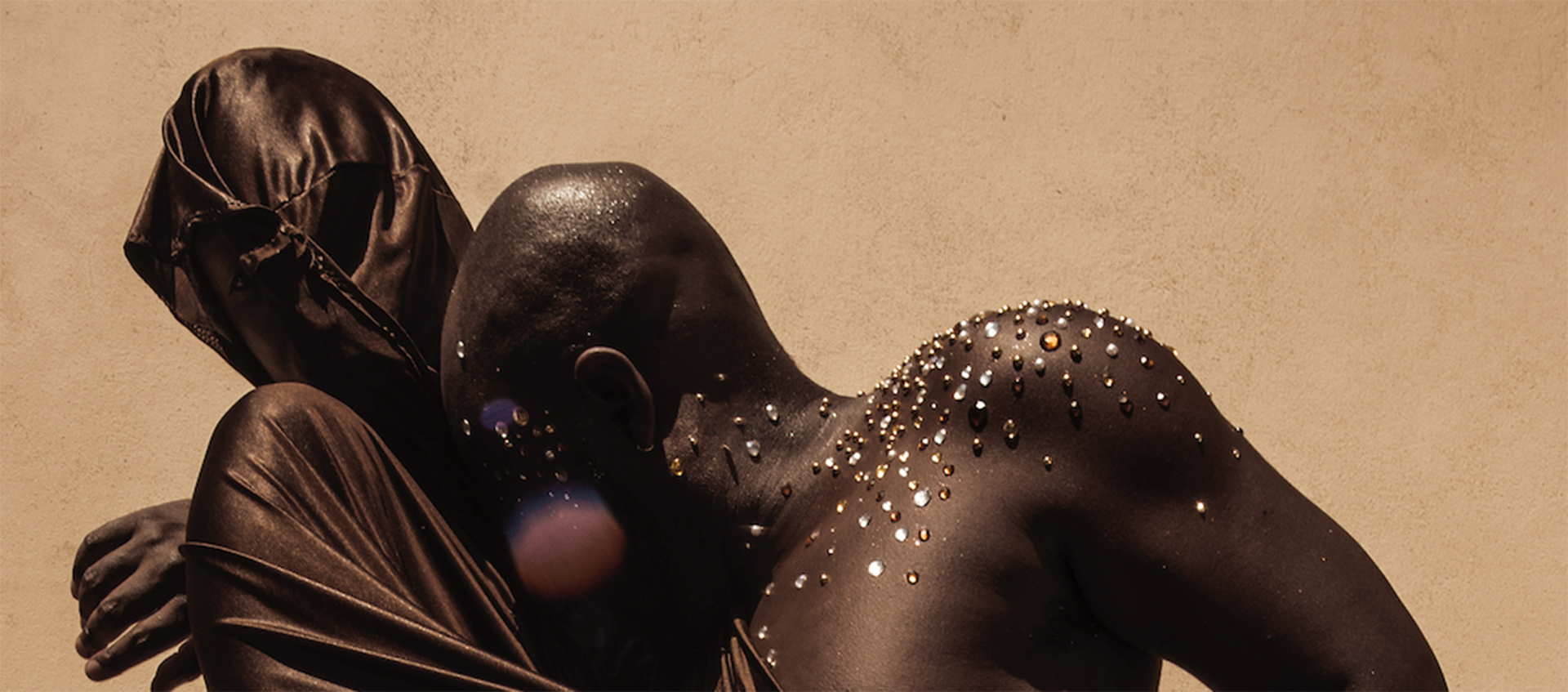 Nile Harris (left) and jaamil olawale kosoko (right) embracing one another. Harris’s face and body are covered with shimmery brown fabric. kosoko—who has dark brown skin and silver, gold, and brown gems on their bare shoulder, neck, and chest—is leaning their bare head on Harris’s chest.
