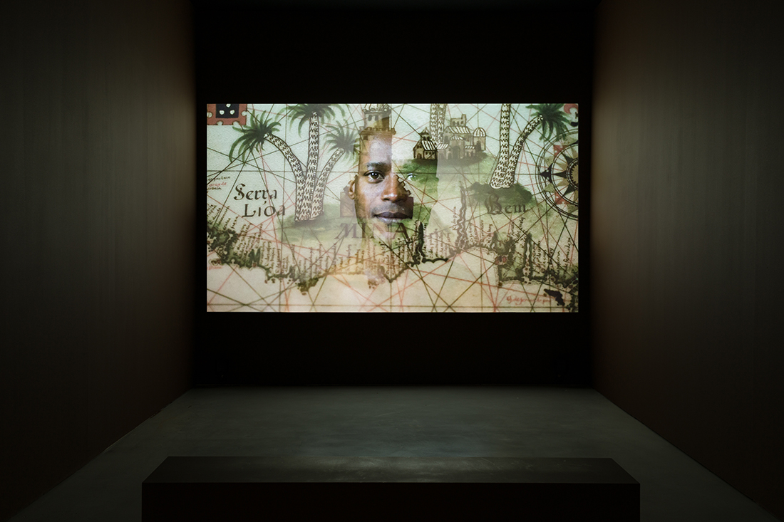 Video projection screen in dark space, featuring a person's face layered atop a drawn historical map with palm trees and a village.