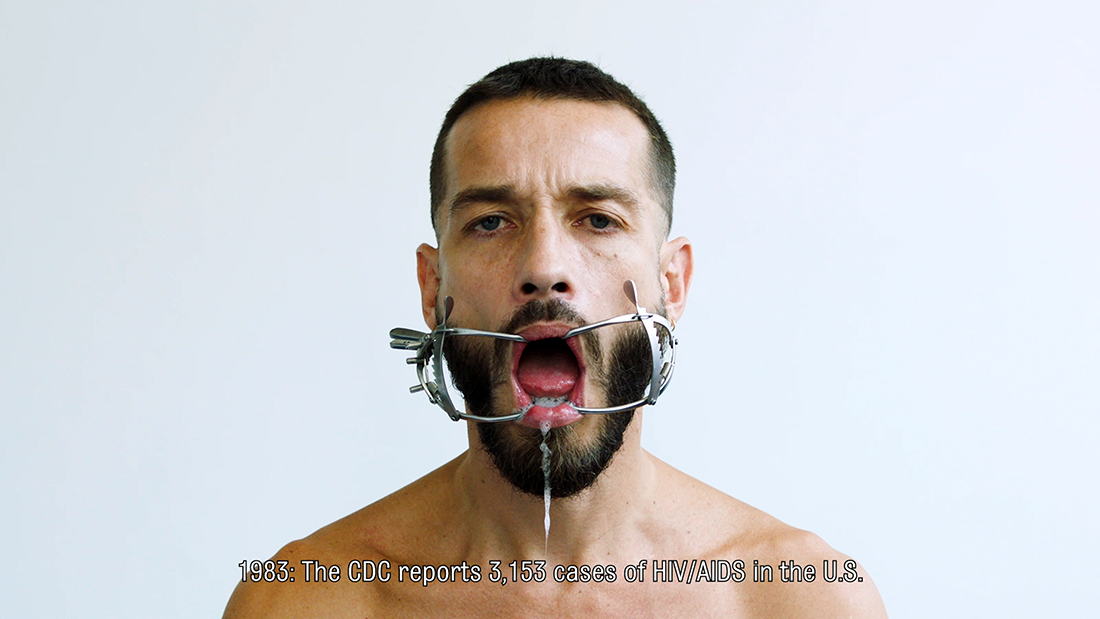 Carlos Motta from his shoulders up; he has a dental gag in his mouth and the following text across his bare chest: "1983: The CDC reports 3,153 cases of HIV/AIDS in the U.S."