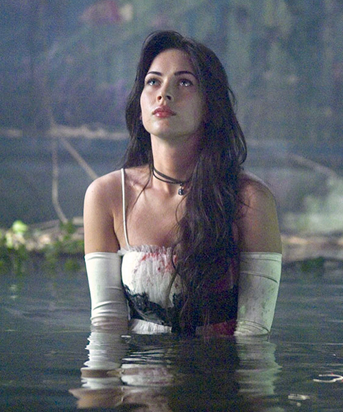Jennifer waist-deep in a pool of water. She is wearing a white dress with a black ribbon and white arm sleeves. Her dress is stained with blood. She is looking up.