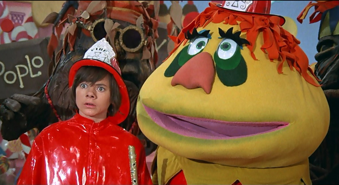 A young boy wearing a red slicker and oversized fireman's hat stares with surprise into the distance and stands next to a brightly yellow colored puppet with a large head.