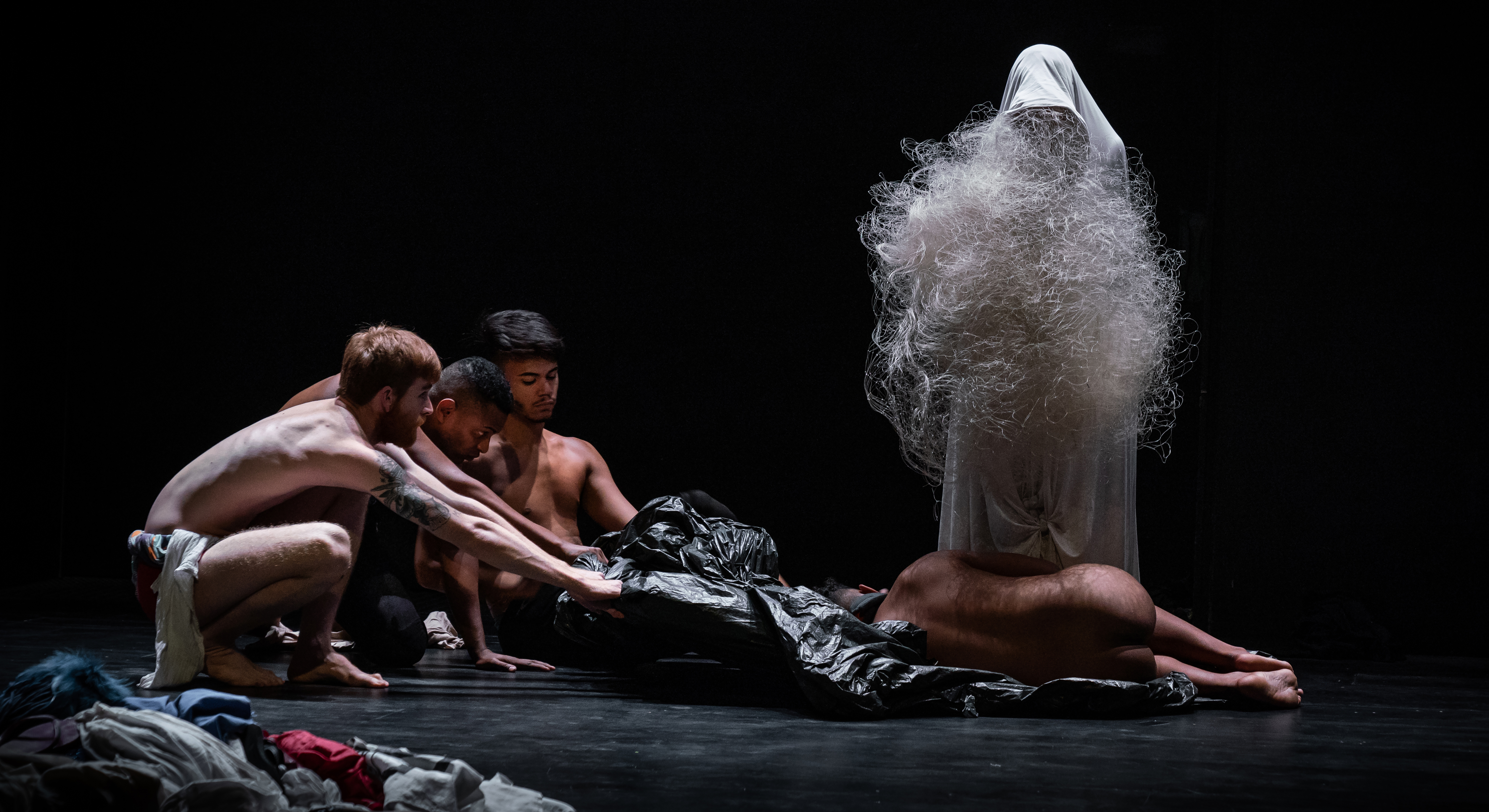 Three shirtless performers pull a large black tarp, on which a nude person is lying in a cradle position. An ominous, white-cloaked figure draped in white, hairy wire looks down at them.