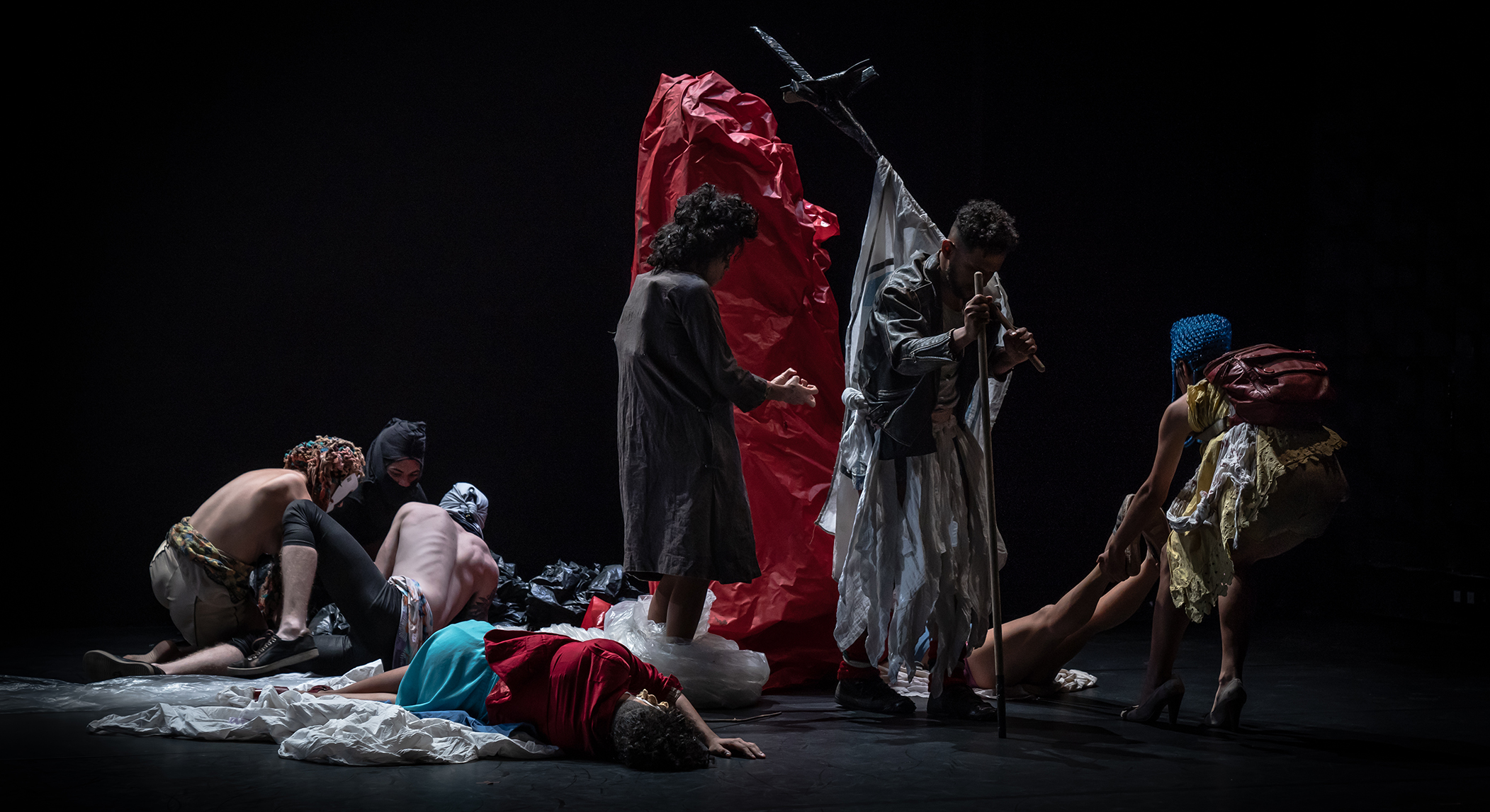 Performers draped in various plastics and fabric are strewn across the stage. Some are lying down, some are walking, and others are being dragged.