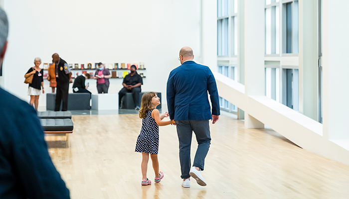 Gallery space featuring a father and child holding hands and walking away from the camera. 
