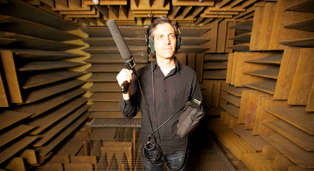 Sam Green wears a black button down, long sleeve shirt untucked atop dark blue jeans. He has close cropped dark hair. He is inside a sound booth and holds a long microphone and wears headphones.