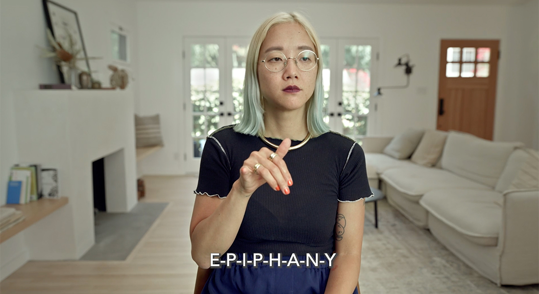 Christine Sun Kim has blond hair with light blue highlights that touches her shoulders. She wears thin, round eyeglasses and sit in a room with a white couch and fireplace with photos and other items on the mantle. Kim is speaking in sign language, and the subtitle word reads "E-P-I-P-H-A-N-Y."