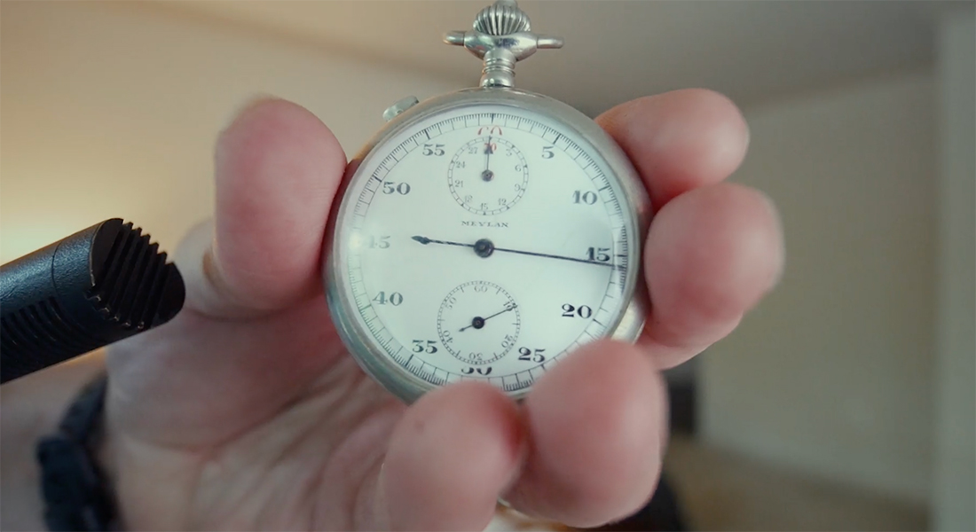 An image of a round stopwatch being gripped by four fingers and a thumb. A small microphone is held just next to it recording its sound.