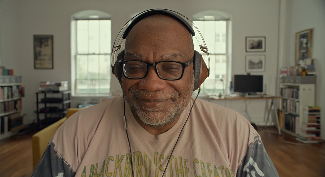 Fred Moten sits with his eyes closed and a slight smile on his lips. He wears black eyeglasses with square frames. He is in what appears to be an office with a desk behind him, upon which sits a computer. Bookshelves are on both sides of the room.