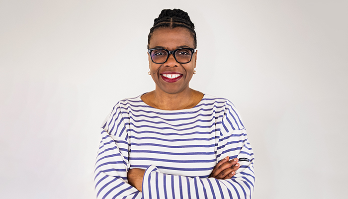 Waist-up photo of Gaëtane Verna. She is a Black woman with braided hair parted at the center and pulled back into a tight bun and black-rimmed glasses. She's wearing small dangling earrings, berry red lipstick, and a long-sleeve, blue and white-striped top. Her arms are folded and she's facing the camera with a warm smile.