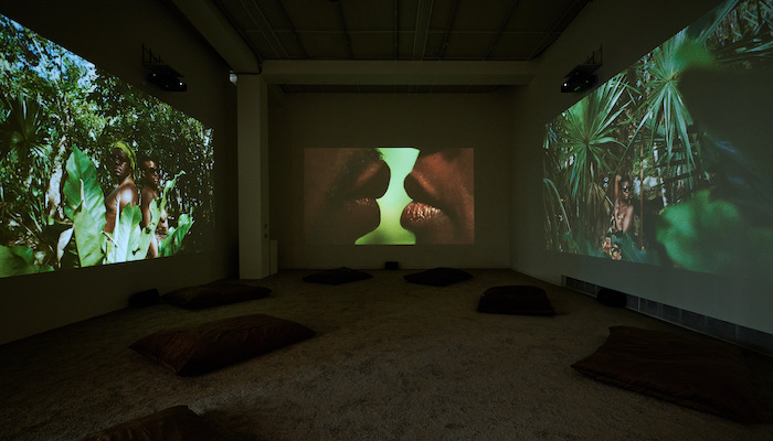 Gallery room with brown pillows scattered throughout the carpeted floor and projectors on the back and side walls. The projector in the back features an up-close shot of two Black people whose lips are separated by a small space, where blurred greenery can be seen in the background. The person on the left has dark hair above their lip, and the person on the right has glossy lips. The side-wall projectors feature three Black people surrounded by green foliage. 