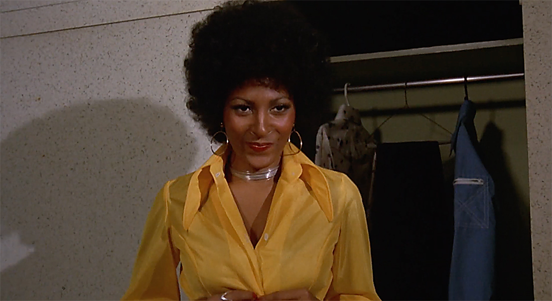 Pam Grier from the waist up in front of a closet. She smirks and wears a yellow button-down shirt. Her hair is styled in an afro.