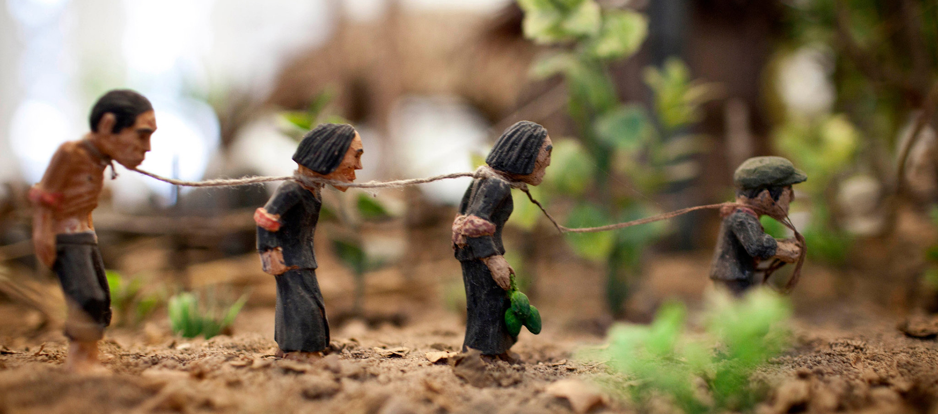 Four clay figurines linked by rope around their necks stand in a line on a dirt path 