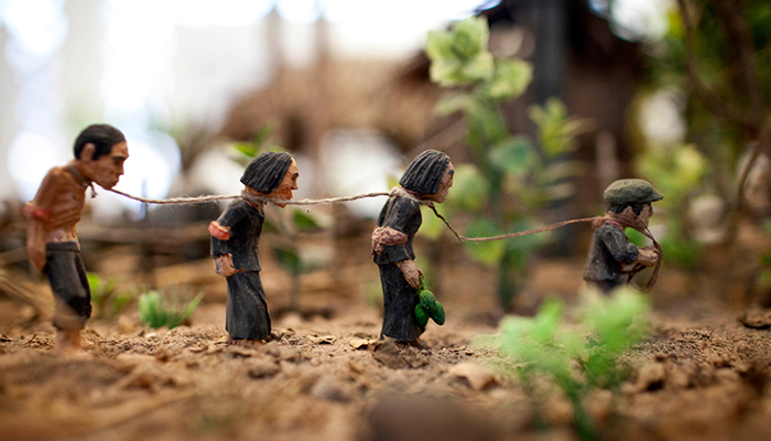 Four clay figurines linked by rope around their necks stand in a line on a dirt path 